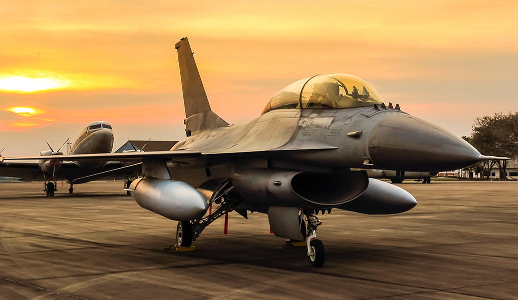 A military fighter jet is parked on the tarmac during sunset, with another aircraft in the background under a warm, golden sky.