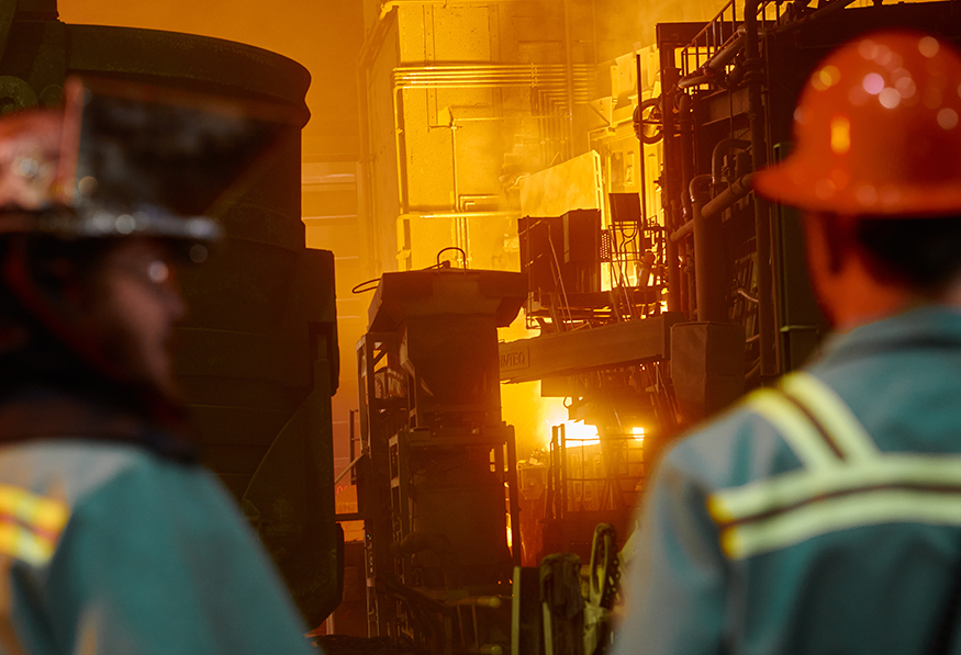 Two people in hard hats and safety gear observe industrial operations with glowing, molten metal, intense heat, and heavy machinery, indicating a steel mill or foundry.