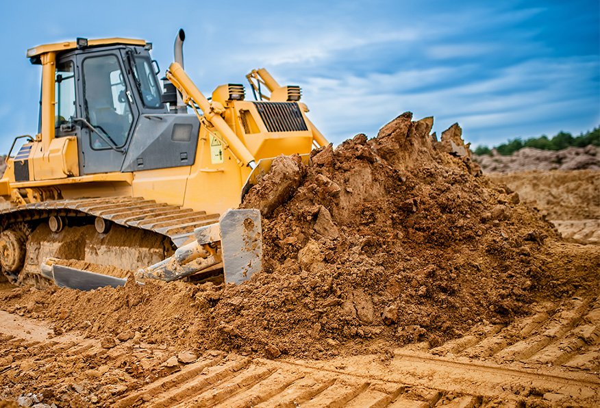 A yellow bulldozer moves earth at a construction site, with its blade pushing a mound of dirt, and track marks imprinted into the soil.