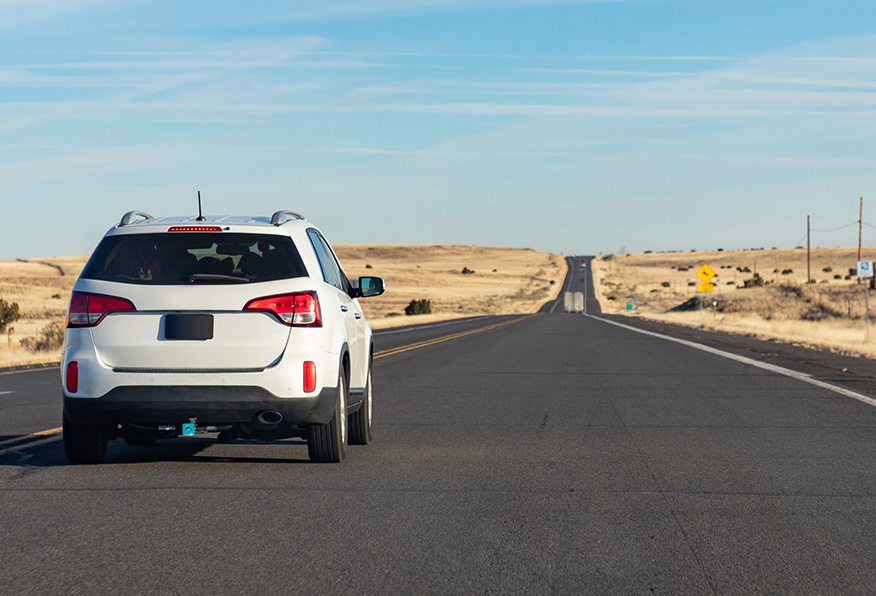 A white SUV is parked on an open road stretching into the distance under a clear blue sky, surrounded by sparse desert landscape.