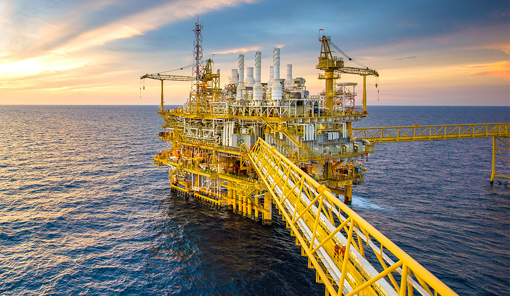 An offshore oil platform stands in the sea during sunset, with extensive infrastructure, multiple levels, and a connecting bridge against a vibrant sky.