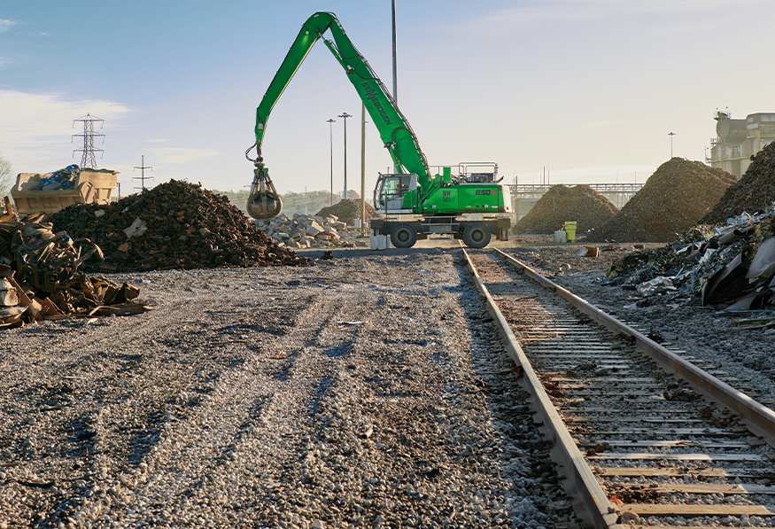 A green excavator with a clamshell bucket is operating in an industrial area with scattered debris, railroad tracks in the foreground, and a clear sky.