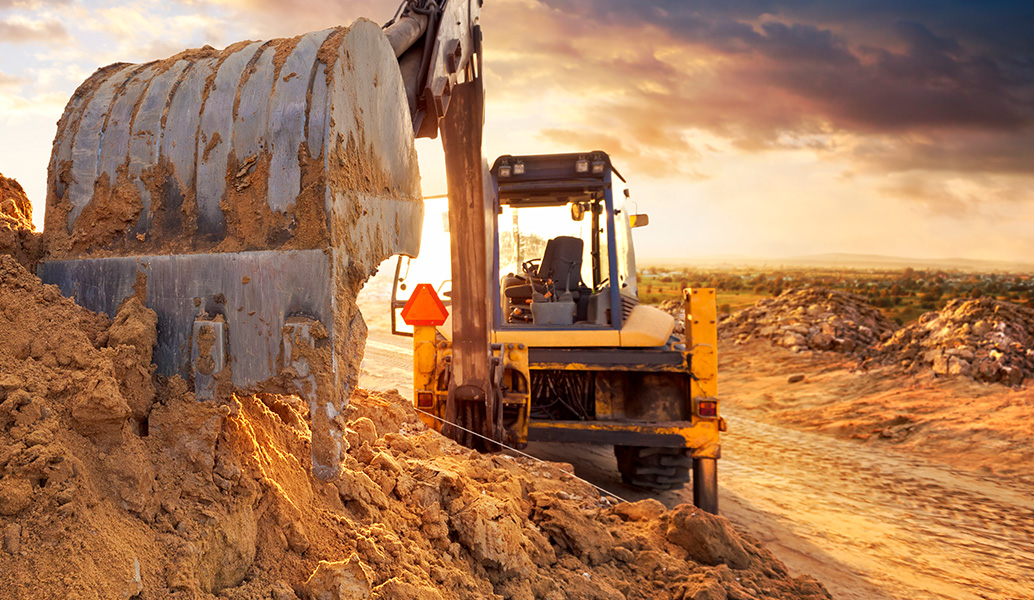 A large yellow excavator operates in a sandy environment under a dramatic sunset sky, its bucket full of earth amid a construction or mining site.