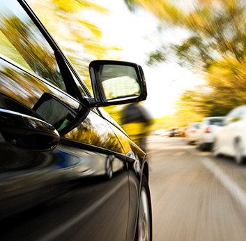 The image shows a black car in motion with a blurred background, depicting movement. The focus is on the side view mirror and the car's silhouette.