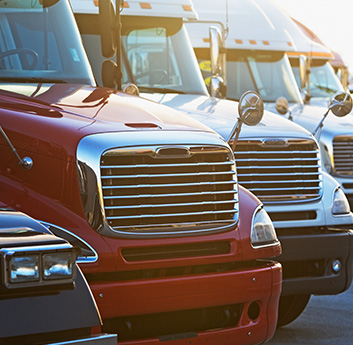A row of parked semi-trucks is shown, featuring prominent grilles and mirrors. The focus is on the red truck in the foreground. The setting appears to be a truck stop or parking lot.