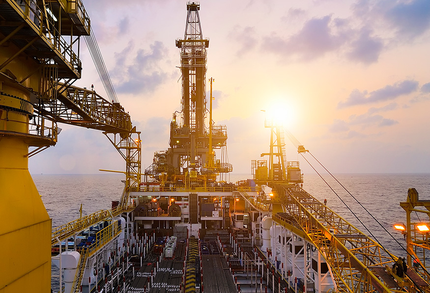 The image shows an offshore oil rig during sunset. The platform is complex with equipment and machinery, and the ocean extends to the horizon.