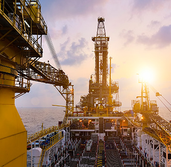 The image shows an offshore oil drilling platform at sunset with intricate machinery, a visible derrick, and the ocean in the background.
