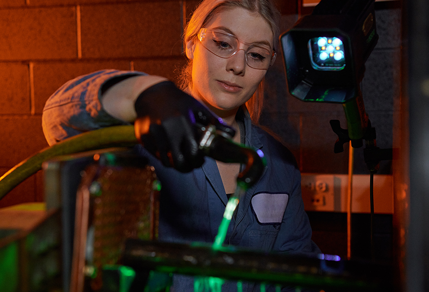 A person is working with machinery in a dimly lit setting, wearing protective glasses and focused on adjusting a component with a green light source nearby.