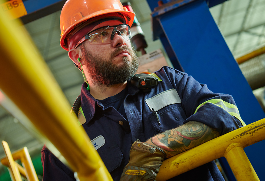 A person in industrial workwear and a hard hat looks thoughtfully off-camera, with safety glasses on their head, standing by yellow railings, tattoo visible on arm.