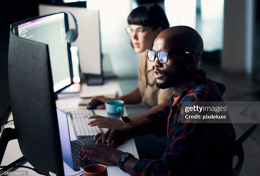 Two people are working in a dimly lit office, focusing intently on computer monitors. The person in the foreground is wearing glasses and a beanie.