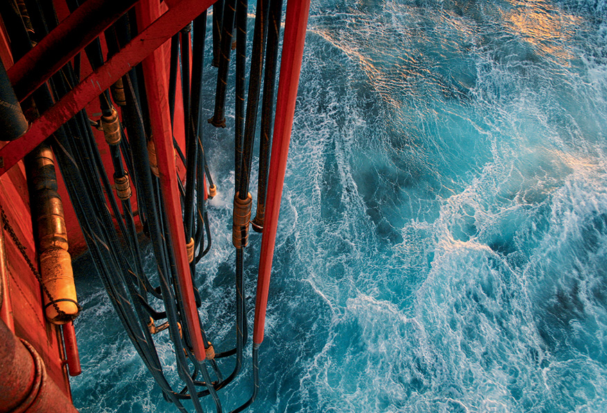 The image shows a high-angle view of turbulent blue ocean waters next to the red steel structure of a ship or oil platform, with pipes and railings.