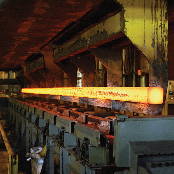 This image shows a glowing, red-hot metal bar on a conveyor inside an industrial setting, likely a steel mill or forge, with visible machinery and structures.