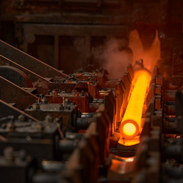 The image shows a glowing hot metal piece on an industrial production line, surrounded by machinery, with steam and heat indicating an intense manufacturing process.