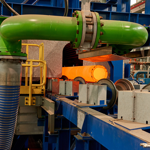 An industrial setting with green pipes, blue steel structures, and an orange cylindrical object amidst machinery, likely part of a manufacturing or processing plant.