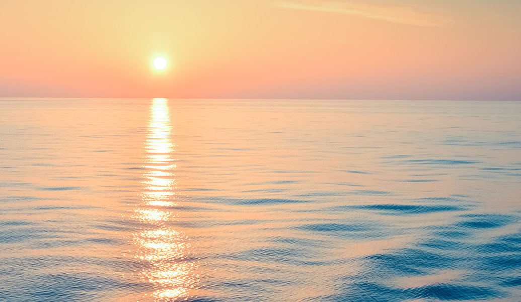 This image features a tranquil sunset with a pastel sky above a calm sea. The sun's reflection creates a shimmering path across the water's surface.