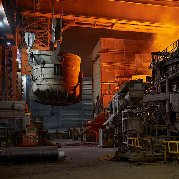 An industrial steel foundry with intense orange glow. A large ladle crane transports molten metal. Heavy machinery surrounds the area, signifying robust manufacturing activity.