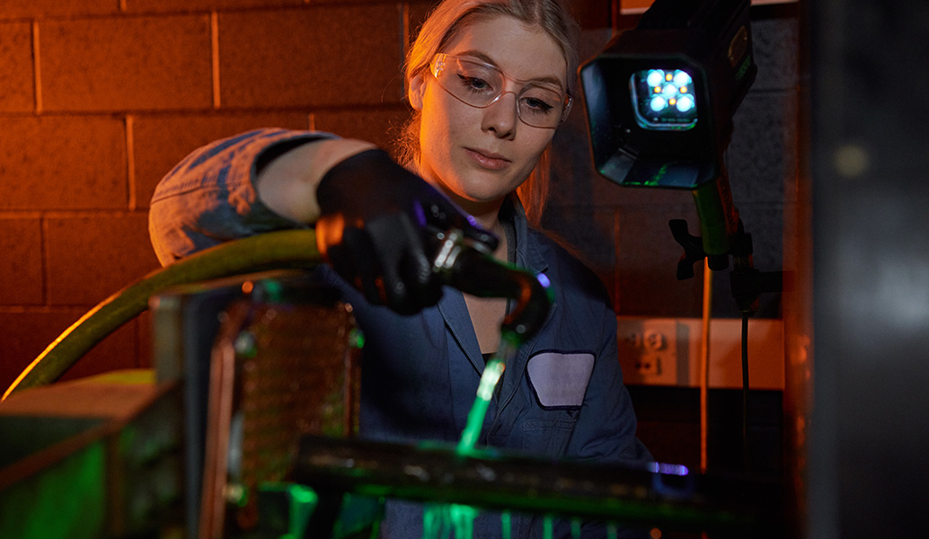 A person in safety glasses is working with machinery in a dimly lit room, focusing intently on their task. Industrial equipment with green light is visible.