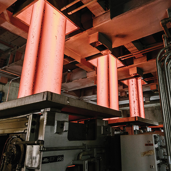 This image shows glowing red-hot steel slabs inside an industrial facility, indicative of a steel mill or metallurgical plant, with heavy machinery around.