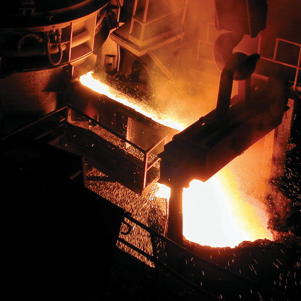 An industrial setting with molten metal being poured from a large furnace into a mold, creating bright, fiery sparks, indicative of a steel mill or foundry.