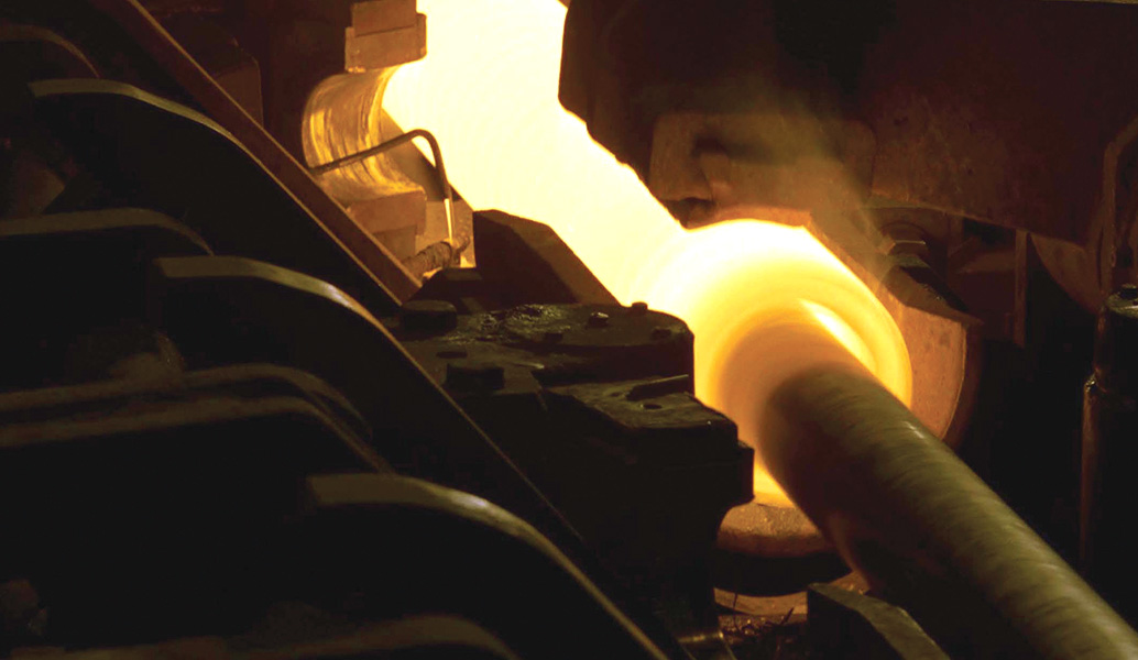 A person works with glowing hot metal in an industrial setting, possibly a forge, amid heavy machinery, showcasing a manufacturing or metalwork process.