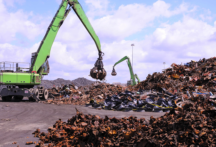 Two large green cranes with grapples are sorting through a vast pile of metal scrap in a recycling facility under a cloudy sky.