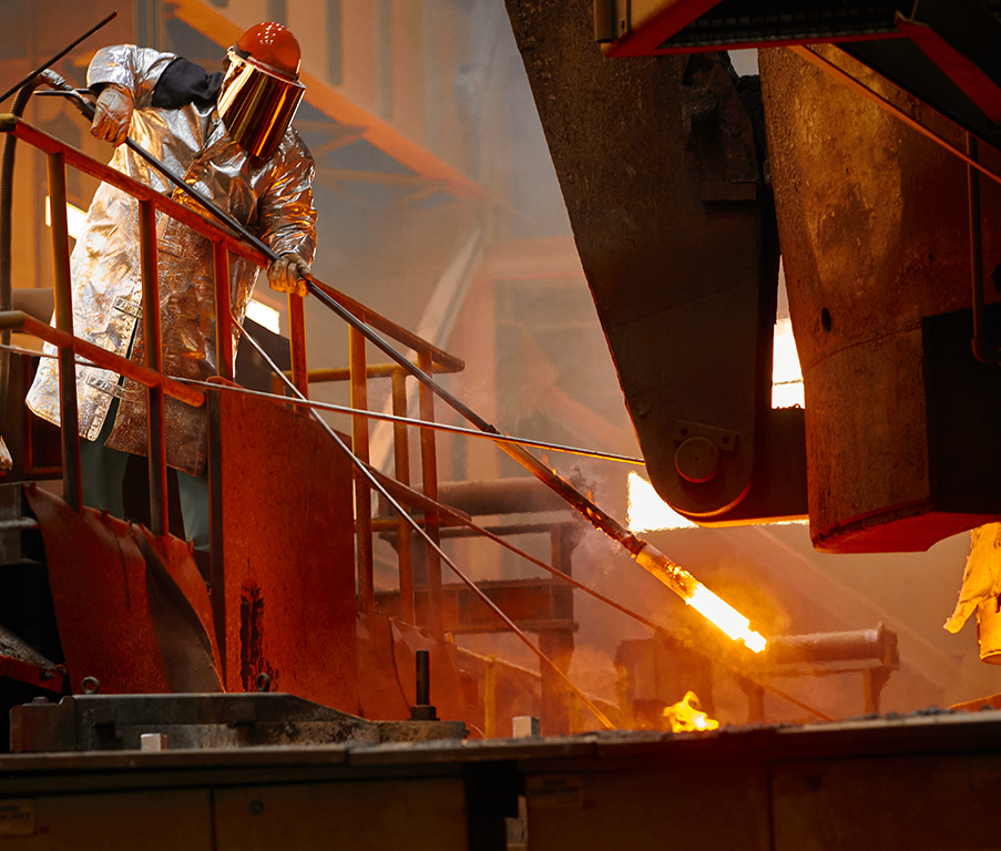 A person in protective gear works with molten metal in an industrial facility, standing beside intense heat and vibrant orange flames.