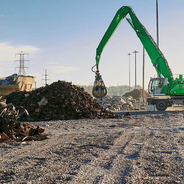 A green excavator with a grapple attachment works at a scrapyard, moving a mix of metal and debris. A clear sky and power lines are visible.