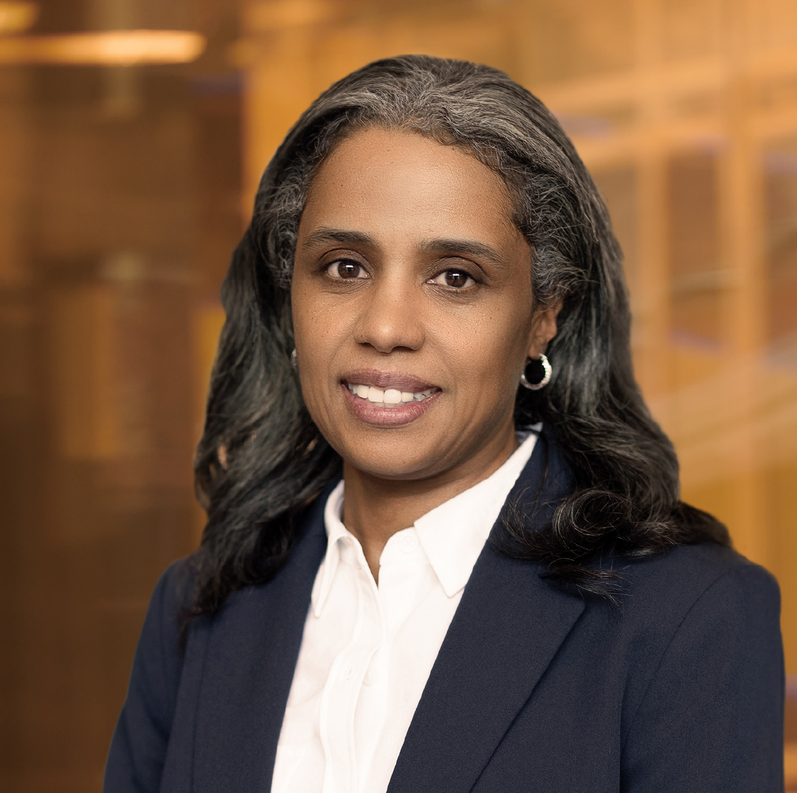 A professional-looking person in a gray blazer and polka dot shirt, smiling, with long hair, set against a blurred indoor background.